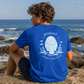 A man wearing a royal blue t-shirt sits on a rock by the ocean, looking out at the water. The back of the t-shirt features a light blue graphic of a manatee surrounded by bubbles, fish, and coral, with the words 'protect what you love' written in a light blue font.