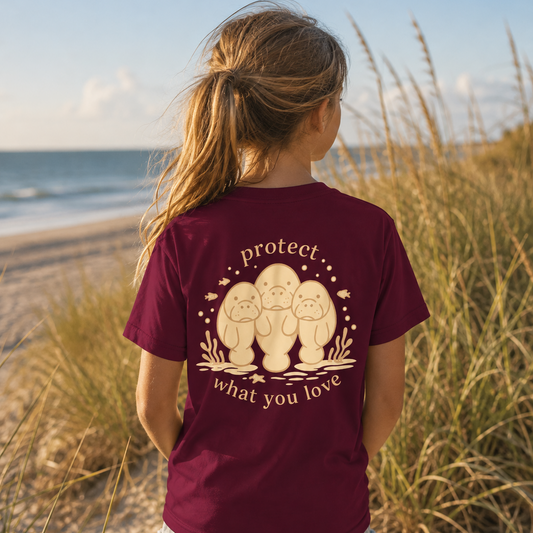 A woman standing on a beach dune wearing a maroon t-shirt with a cream graphic of three pugs and the text 'protect what you love', seen from behind.