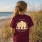A woman standing on a beach dune wearing a maroon t-shirt with a cream graphic of three pugs and the text 'protect what you love', seen from behind.