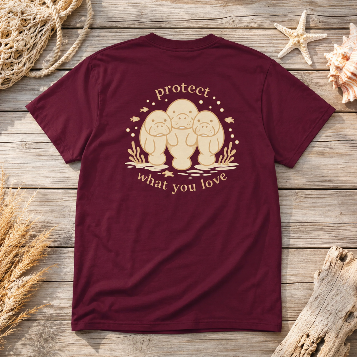A maroon short-sleeve t-shirt laid flat on a rustic wooden surface, decorated with seashells, driftwood, and rope. The back of the shirt features a beige graphic of three baby bonnetheads with the words 'protect what you love' written around them.