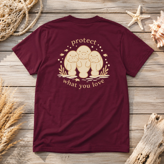 A maroon short-sleeve t-shirt laid flat on a rustic wooden surface, decorated with seashells, driftwood, and rope. The back of the shirt features a beige graphic of three baby bonnetheads with the words 'protect what you love' written around them.