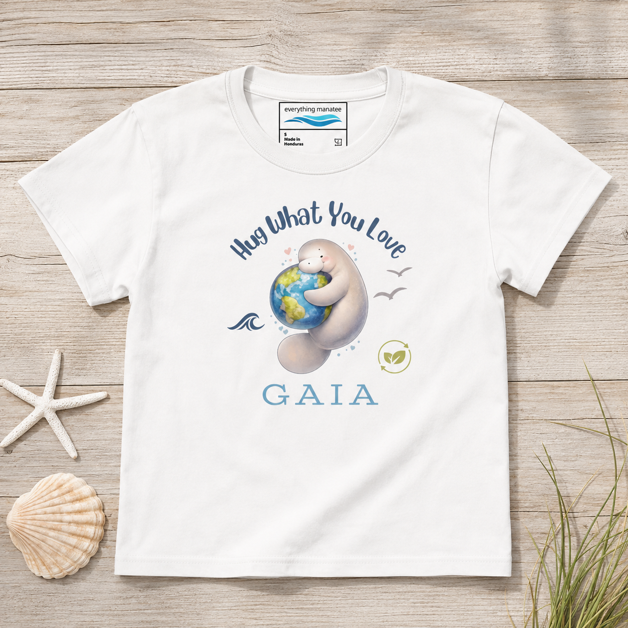 A white short-sleeve t-shirt with a custom graphic print of a manatee hugging a globe and the text 'Hug What You Love GAIA'. The shirt is displayed flat on a light wood background with decorative seashells and grass.