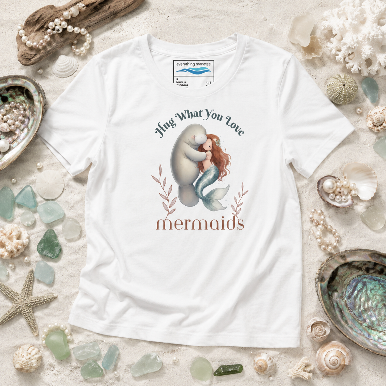 A white graphic t-shirt featuring a mermaid hugging a man with the text 'Hug What You Love mermaids' printed on the front. The shirt is displayed in a flat-lay arrangement on a sandy background decorated with seashells, sea glass, and driftwood.