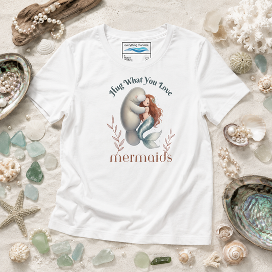 A white graphic t-shirt featuring a mermaid hugging a man with the text 'Hug What You Love mermaids' printed on the front. The shirt is displayed in a flat-lay arrangement on a sandy background decorated with seashells, sea glass, and driftwood.