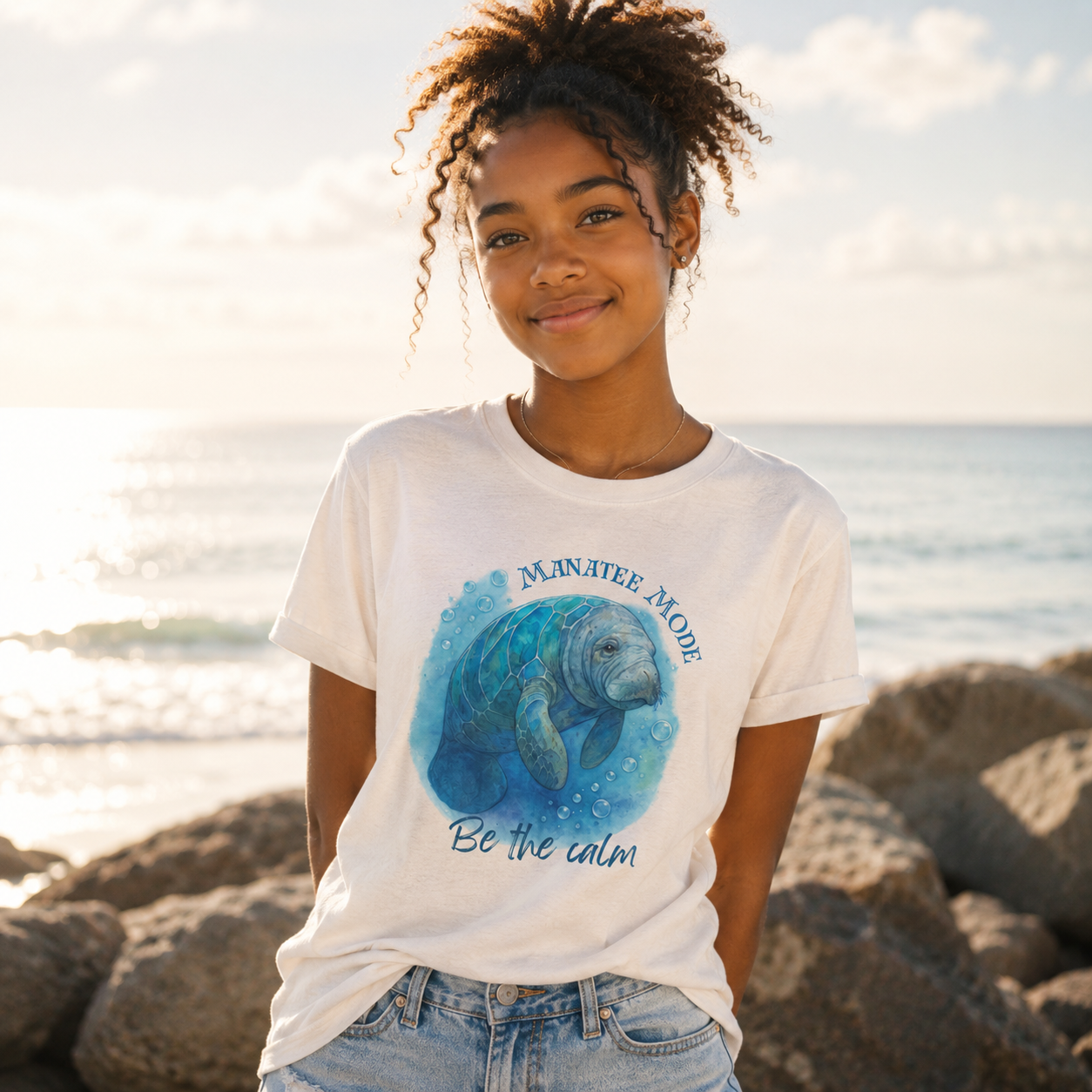 A young woman with curly hair stands on rocks by the ocean wearing a white graphic t-shirt with a blue manatee illustration and the text 'MANATEE MODE Be the calm'.
