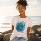 A young woman with curly hair stands on rocks by the ocean wearing a white graphic t-shirt with a blue manatee illustration and the text 'MANATEE MODE Be the calm'.