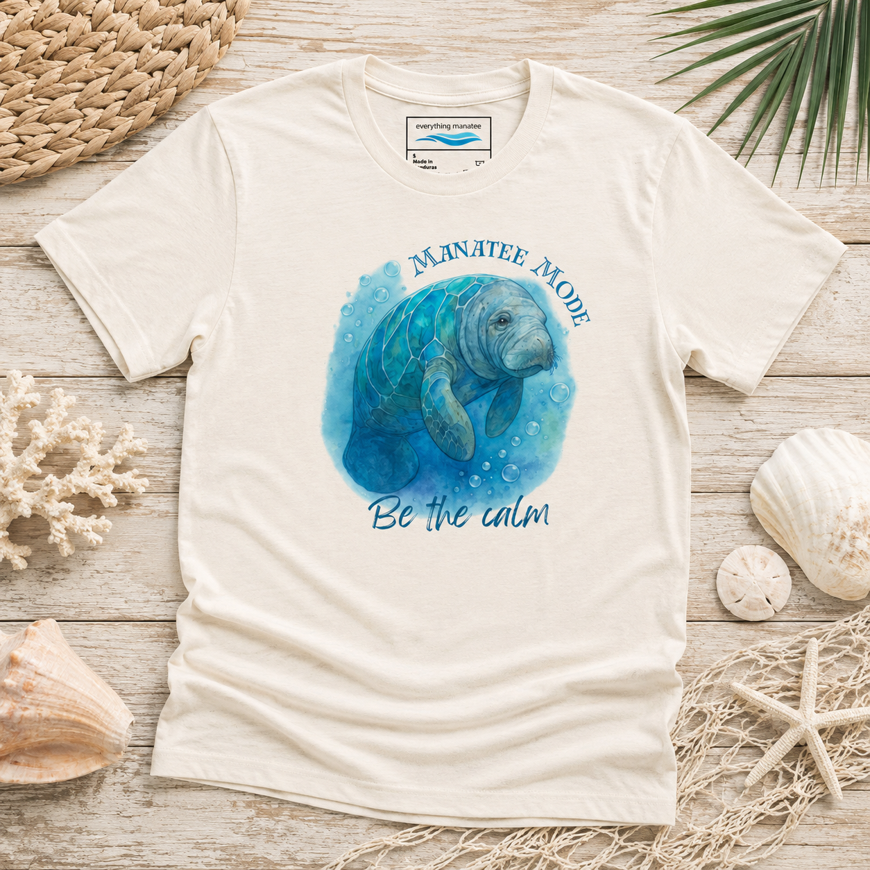 A cream-colored short-sleeve t-shirt with a blue watercolor manatee graphic and the text 'MANATEE MODE Be the calm' printed on the front. The shirt is styled in a flat-lay arrangement on a light wood surface with seashells, a woven mat, and palm leaves.