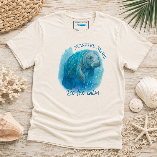 A cream-colored short-sleeve t-shirt with a blue watercolor manatee graphic and the text 'MANATEE MODE Be the calm' printed on the front. The shirt is styled in a flat-lay arrangement on a light wood surface with seashells, a woven mat, and palm leaves.