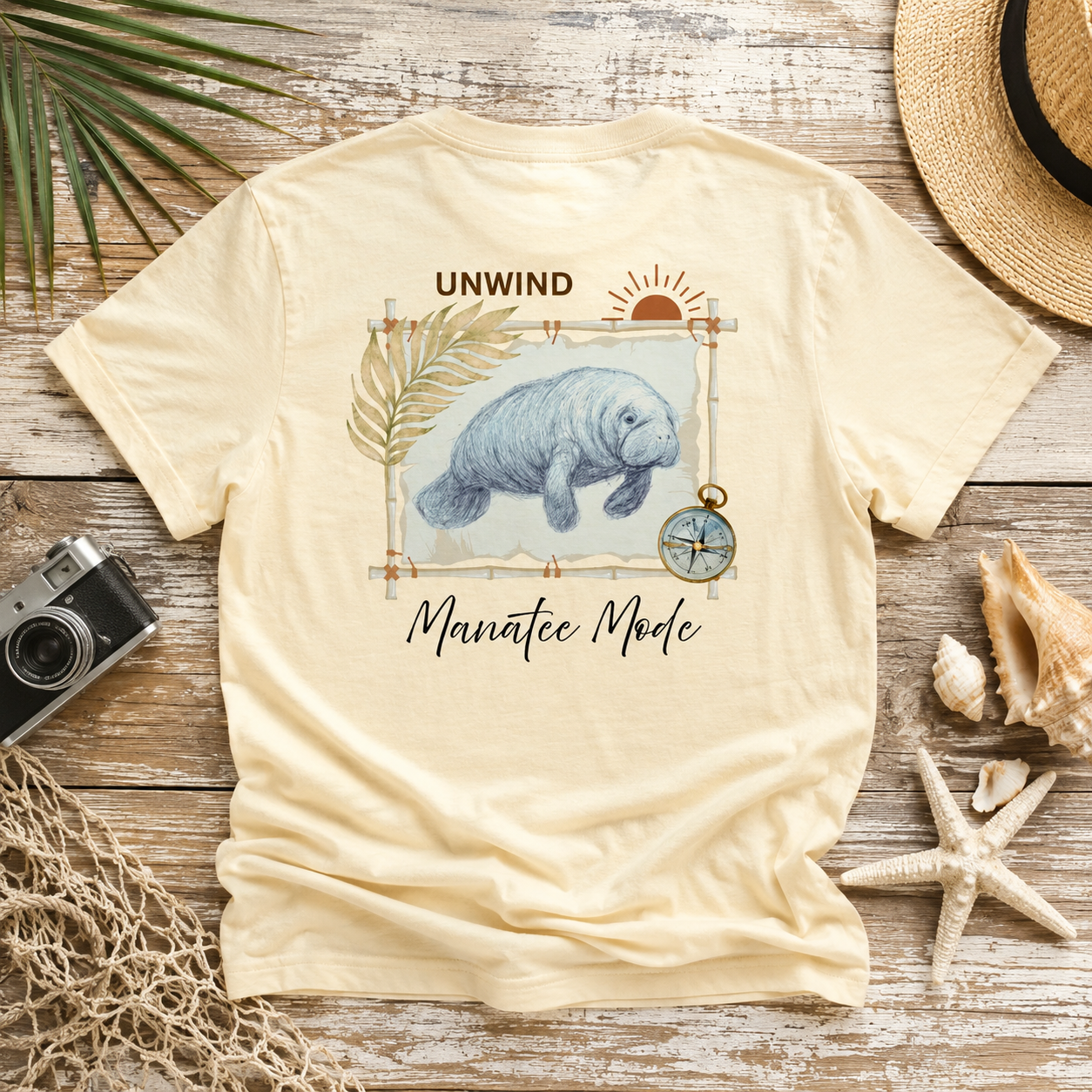A cream-colored t-shirt with a manatee graphic and the text 'UNWIND Manatee Mode', styled in a flat-lay on a rustic wooden background with a straw hat, seashells, a camera, and netting.