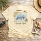 A cream-colored t-shirt with a manatee graphic and the text 'UNWIND Manatee Mode', styled in a flat-lay on a rustic wooden background with a straw hat, seashells, a camera, and netting.