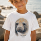 A young boy wearing a white t-shirt with a colorful watercolor-style graphic of a manatee face and the text 'The Gentle Giant', set against a blurred rocky beach background.