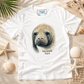A white short-sleeve graphic T-shirt laid flat on a sandy beach-style surface with seashells, starfish, driftwood, and palm leaves. The shirt features a large printed illustration of a manatee face with the text 'The Gentle Giant' beneath it.