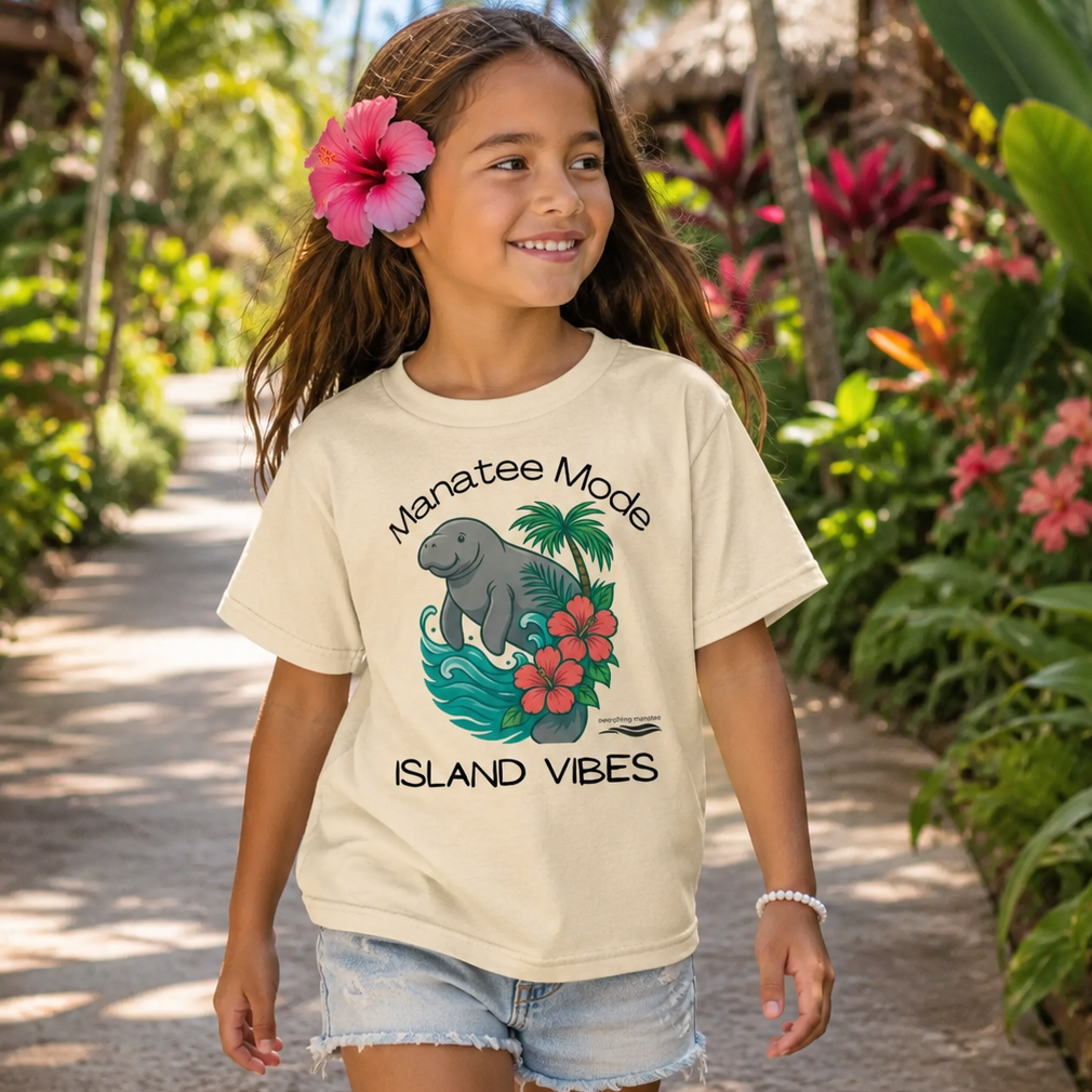 A young girl with a pink hibiscus flower in her hair walks down a tropical garden path wearing a cream-colored graphic t-shirt that reads 'Manatee Mode Island Vibes' with a graphic of an elephant, palm tree, and red flowers.
