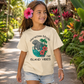 A young girl with a pink hibiscus flower in her hair walks down a tropical garden path wearing a cream-colored graphic t-shirt that reads 'Manatee Mode Island Vibes' with a graphic of an elephant, palm tree, and red flowers.