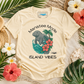 A cream-colored graphic t-shirt laid flat on a sandy surface surrounded by tropical leaves, hibiscus flowers, and woven straw accessories. The shirt features a graphic of a gray manatee on a wave with pink hibiscus flowers and the text 'Manatee Mode ISLAND VIBES'.