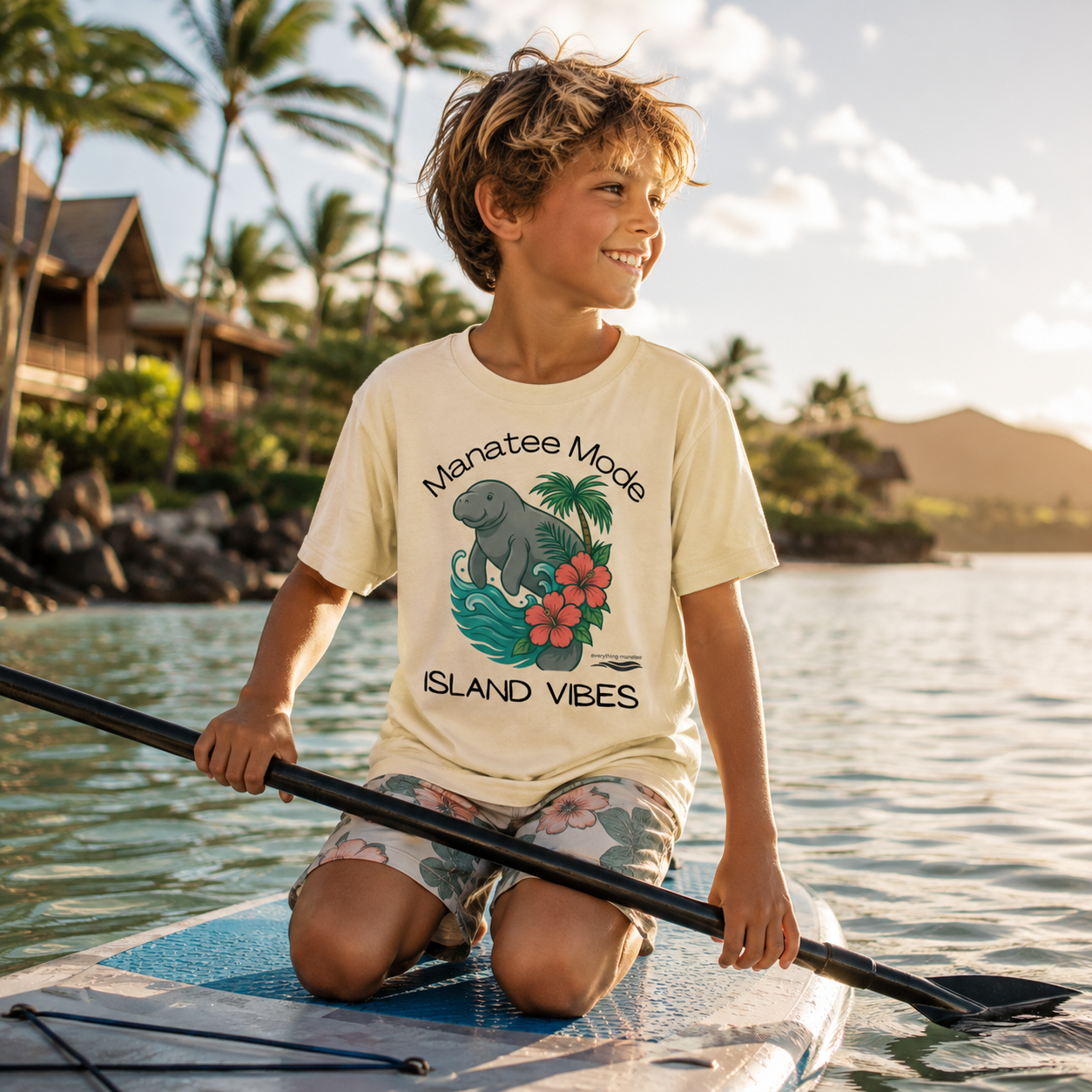 A smiling boy kneeling on a paddleboard in the water at a tropical beach, wearing a cream graphic T-shirt that reads "Manatee Mode Island Vibes" with a large elephant and floral design.