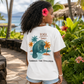 A woman stands outdoors on a tropical path wearing a white graphic T-shirt with a manatee and palm tree design and the words 'BEACH time' and 'EMBRACE THE STILLNESS.'.