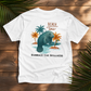 A white short-sleeve graphic t-shirt laid flat on a rustic wooden background, surrounded by green palm leaves. The graphic features a blue-gray manatee in water with orange palm trees and the text 'BEACH time' and 'EMBRACE THE STILLNESS'.