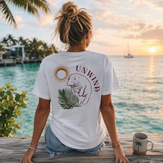 A woman sits on a wooden dock at the ocean during sunset, viewed from behind. She is wearing a white graphic T-shirt with a manatee, palm leaves, and the word 'UNWIND' printed on the back, paired with denim shorts.