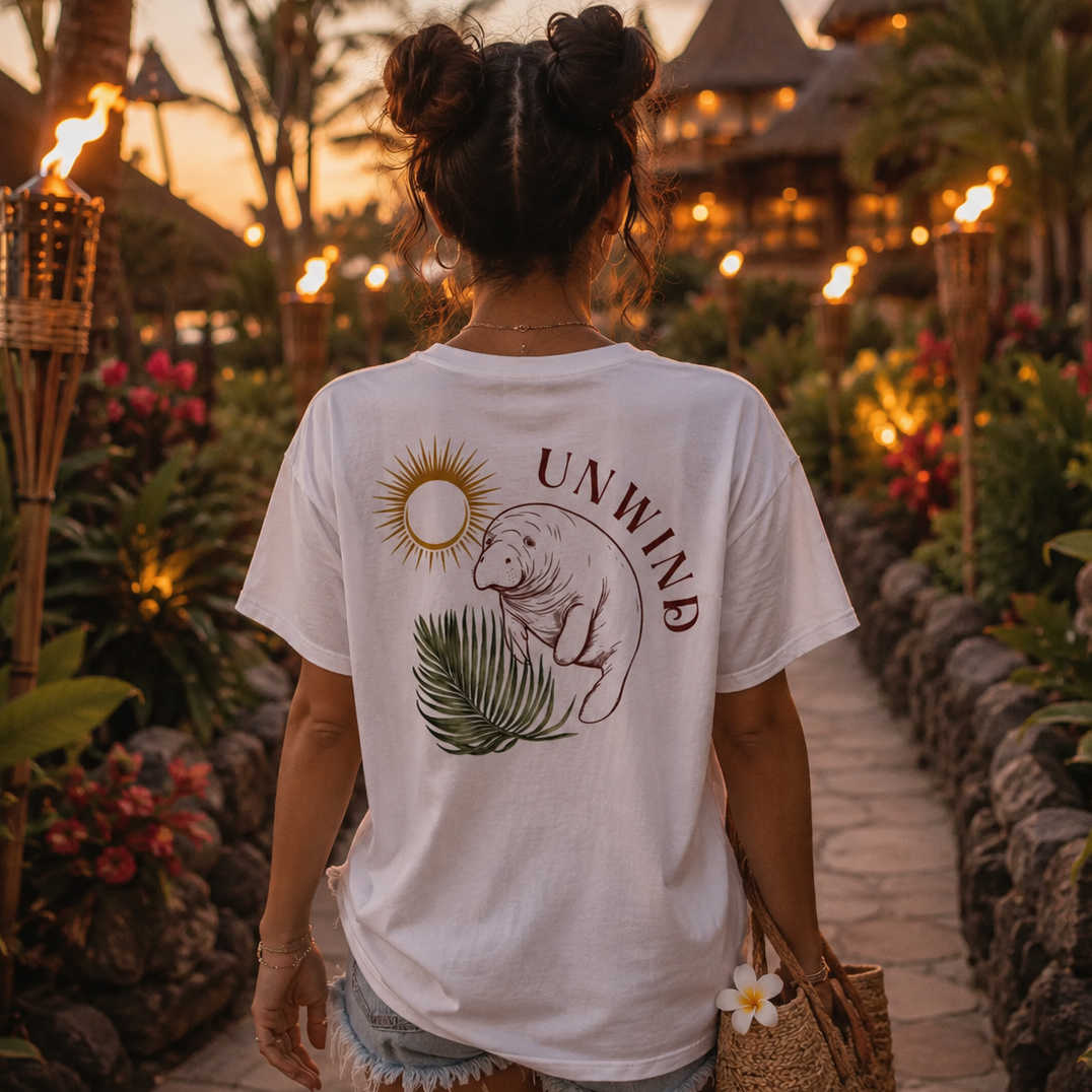 A woman viewed from behind wearing a white graphic t-shirt with a manatee and sun design, standing on a tropical resort path at sunset.