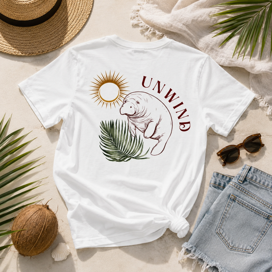 A white graphic t-shirt featuring a manatee, sun, and palm leaf design with the word 'UNWIND' in maroon, styled in a flat lay with a straw hat, denim shorts, sunglasses, a coconut, and palm leaves on a light beige background.