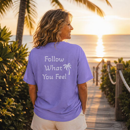 A woman stands on a boardwalk at sunset facing the ocean, wearing a lavender-purple t-shirt with the white text 'Follow What You Feel' and a small palm tree graphic on the back.