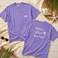 Two purple short-sleeve T-shirts arranged on a light wood surface with beach-themed props. The front shirt shows a small white polar bear graphic on the chest pocket, and the back shirt has white text reading 'Follow What You Feel' with a palm tree graphic.