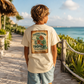 A child viewed from behind wearing a cream-colored t-shirt with a large tropical turtle graphic on the back, standing on a stone walkway overlooking a tropical ocean.