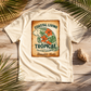 A cream-colored short-sleeve t-shirt laid flat on a rustic wooden surface, surrounded by palm leaves and a seashell. The shirt features a large rectangular graphic on the back with a vintage-style tan background, showing a green turtle, orange hibiscus flowers, and the text 'COASTAL LIVING TROPICAL VACATION PARADISE Manatee Mode'.