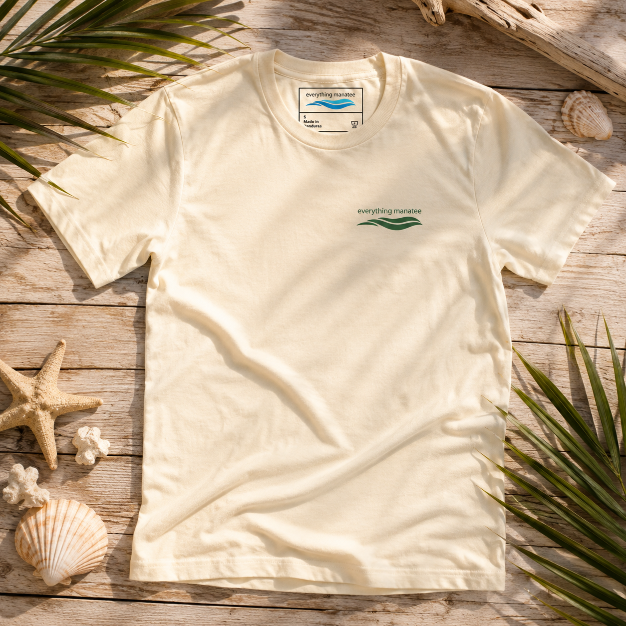An off-white short-sleeve t-shirt with a green wave logo and 'everything marine' text on the left chest, laid flat on a rustic wooden surface surrounded by seashells, a starfish, and palm leaves.