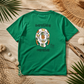 A green short-sleeve t-shirt laid flat on a rustic wooden surface, surrounded by beach-themed props like palm leaves, a woven hat, driftwood, and seashells. The back of the shirt features a graphic of a white turtle shell decorated with pineapples and lemons, with the text 'everything manatee' written above and below the illustration.