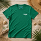 A green short-sleeve t-shirt with a white 'everything manual' logo on the left chest, laid flat on a wooden surface surrounded by beach-themed props like palm leaves, a straw hat, and seashells.