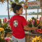 A woman stands at a tropical flower stall wearing a bright red t-shirt with a graphic on the back. The graphic features a white turtle with colorful floral details and the text 'everything manatee'.