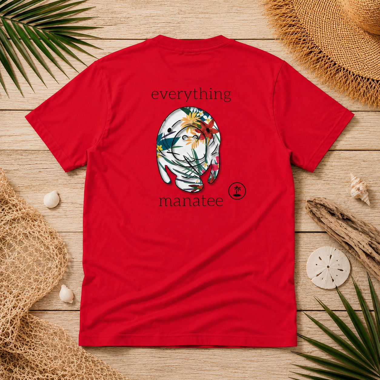A red short-sleeve t-shirt laid flat on a light wood surface, surrounded by beach-themed props including palm leaves, a straw hat, driftwood, and seashells. The shirt features a central graphic of a white cat face decorated with tropical flowers and the words 'everything' above and 'manatee' below.