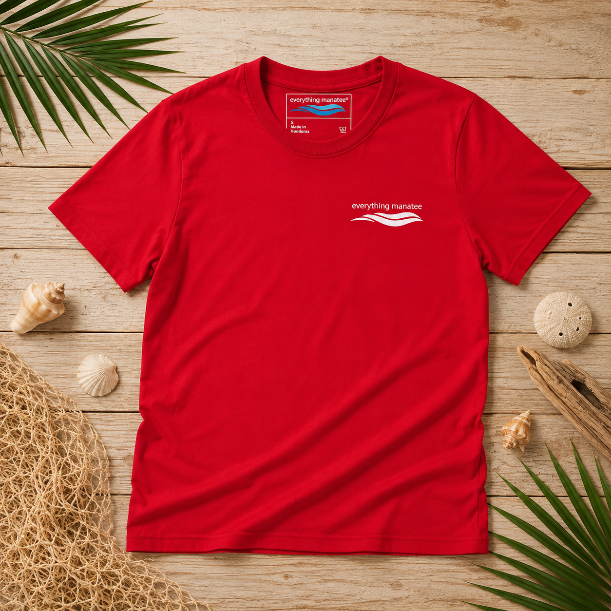A bright red short-sleeve t-shirt with a small white 'everything mandate' logo on the left chest, laid flat on a light wood plank background surrounded by beach-themed props like shells, palm leaves, and a straw hat.