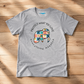 A heather gray short-sleeve t-shirt laid flat on a light wood background. The shirt features a colorful patchwork elephant graphic in the center with the text 'PROTECT WHAT YOU LOVE' arched above it and 'everything manatee' curved below it.