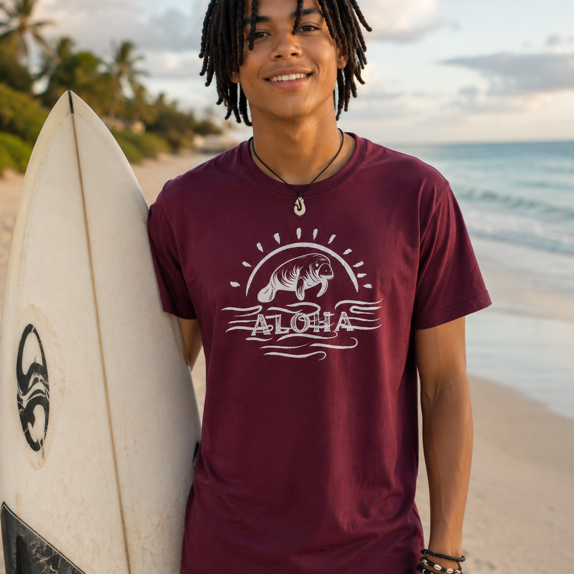 A smiling man with dreadlocks stands on a beach holding a white surfboard, wearing a maroon short-sleeve t-shirt with a white graphic of a bear, waves, and the word 'ALOHA'.
