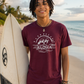 A smiling man with dreadlocks stands on a beach holding a white surfboard, wearing a maroon short-sleeve t-shirt with a white graphic of a bear, waves, and the word 'ALOHA'.