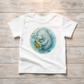 A white toddler short-sleeve t-shirt with a watercolor-style graphic of a blue manatee hugging a green sea turtle inside a circular frame, with the text 'The Great Green Giant' on the chest. The t-shirt is laid flat on a light-colored wooden plank background.