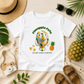 A white short-sleeve graphic T-shirt laid flat on a light wood surface with beach and summer props. The shirt has a cartoon character covered in pineapple slices with the text 'Pineapple Pete' above and 'ISLAND VIBES MANATEE' below, alongside a pineapple graphic.
