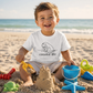 A young child sitting on a sandy beach playing with toy sanders and a bucket, wearing a white t-shirt with a black line art graphic of a seal and the text 'coastal life'.