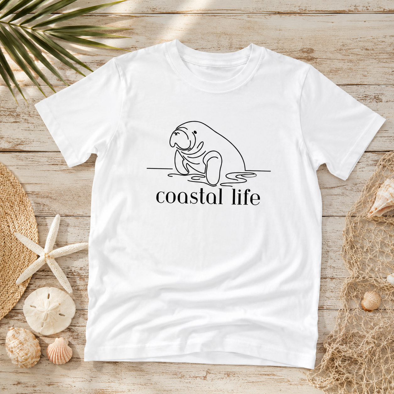 A white short-sleeve T-shirt laid flat on a light wood surface, featuring a black line drawing of a walrus above the words 'coastal life.' The shirt is styled with beach-themed props including shells, a starfish, netting, and palm leaves.