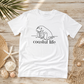 A white short-sleeve T-shirt laid flat on a light wood surface, featuring a black line drawing of a walrus above the words 'coastal life.' The shirt is styled with beach-themed props including shells, a starfish, netting, and palm leaves.