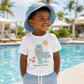 A young child wearing a white graphic t-shirt with a manatee design and the text 'Manatee Mode', paired with a blue bucket hat and light blue pants, standing in front of a swimming pool.