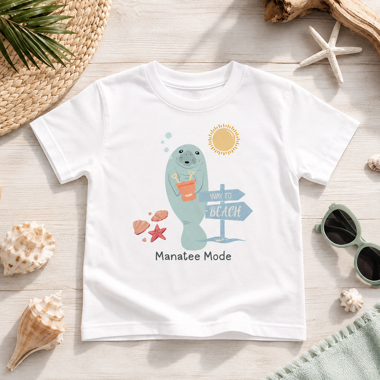 A white short-sleeve T-shirt laid flat on a light wood surface with beach-themed props around it. The shirt has a colorful cartoon manatee graphic holding an orange drink, with a sun, directional sign, shells, and the text 'Manatee Mode' printed below.