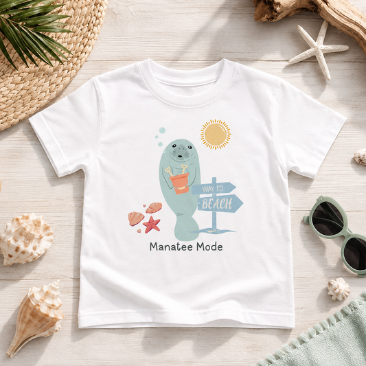 A white short-sleeve T-shirt laid flat on a light wood surface with beach-themed props around it. The shirt has a colorful cartoon manatee graphic holding an orange drink, with a sun, directional sign, shells, and the text 'Manatee Mode' printed below.