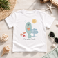 A white short-sleeve T-shirt laid flat on a light wood surface with beach-themed props around it. The shirt has a colorful cartoon manatee graphic holding an orange drink, with a sun, directional sign, shells, and the text 'Manatee Mode' printed below.