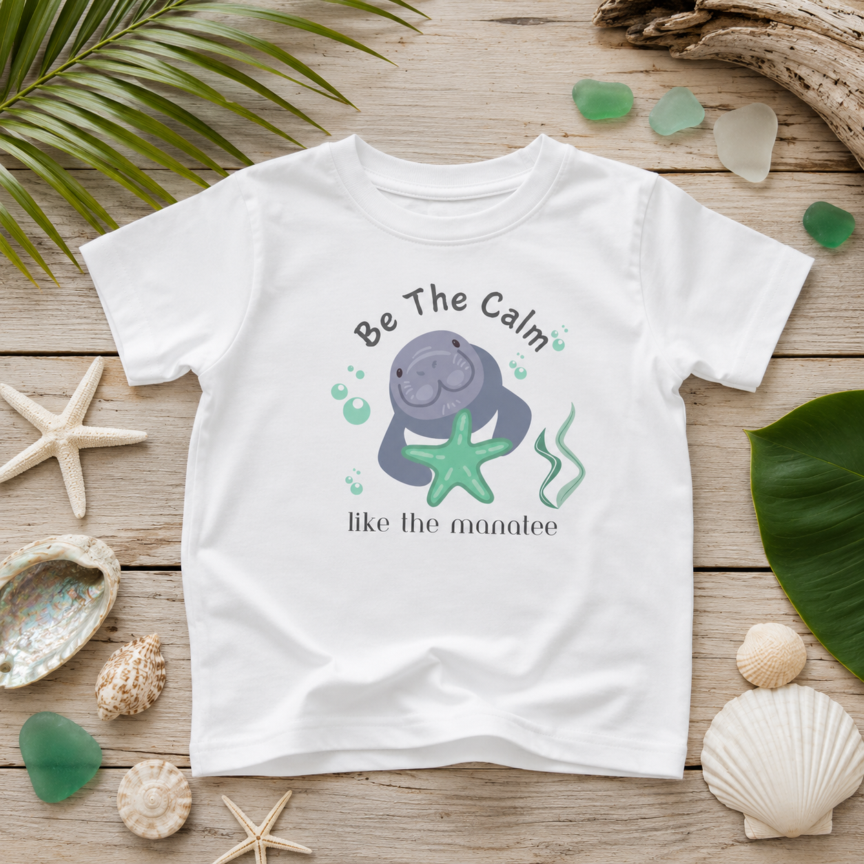 A white kids' t-shirt with a cartoon manatee graphic and the text 'Be The Calm like the manatee', displayed in a flat-lay on a wooden surface surrounded by seashells, sea glass, and palm leaves.