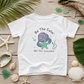 A white kids' t-shirt with a cartoon manatee graphic and the text 'Be The Calm like the manatee', displayed in a flat-lay on a wooden surface surrounded by seashells, sea glass, and palm leaves.