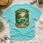 A turquoise short-sleeve t-shirt laid flat on a white wood surface, featuring a large vintage-style graphic on the back with a manatee in a river and the text 'Everything Manatee Florida Native' and 'Sanctuary Waters.' Surrounding props include a straw hat, seashells, a starfish, plant leaves, and a small succulent.